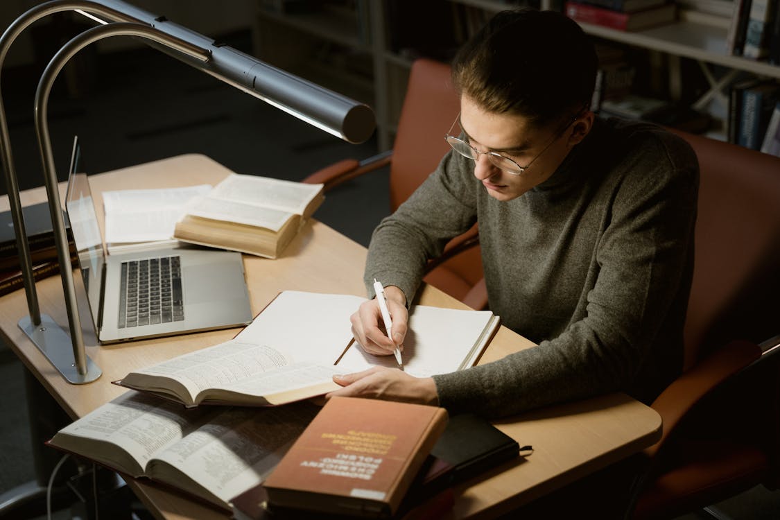 Law student with glasses taking notes surrounded by open casebooks at a library desk